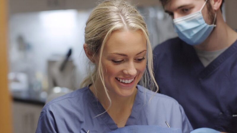 A smiling dental professional in scrubs with a colleague wearing a mask in the background