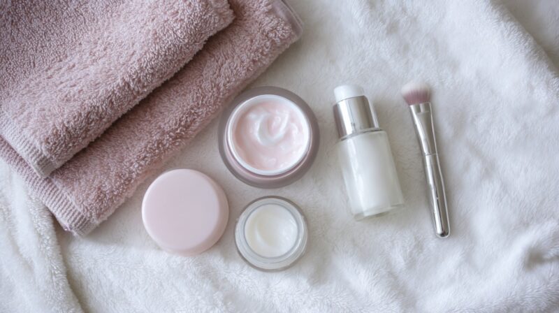 Flat lay of skincare products including creams, lotion, makeup brush, and folded towels on a soft fabric